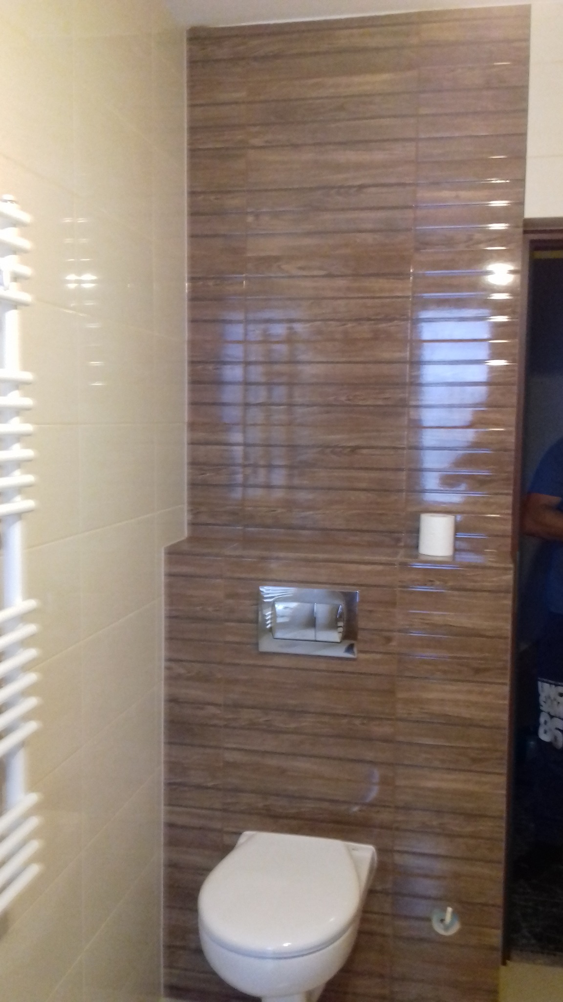 Nowoczesna łazienka z wiszącą toaletą, brązowe płytki imitujące drewno na ścianie za toaletą, obok beżowe płytki, biały grzejnik drabinkowy po lewej stronie.