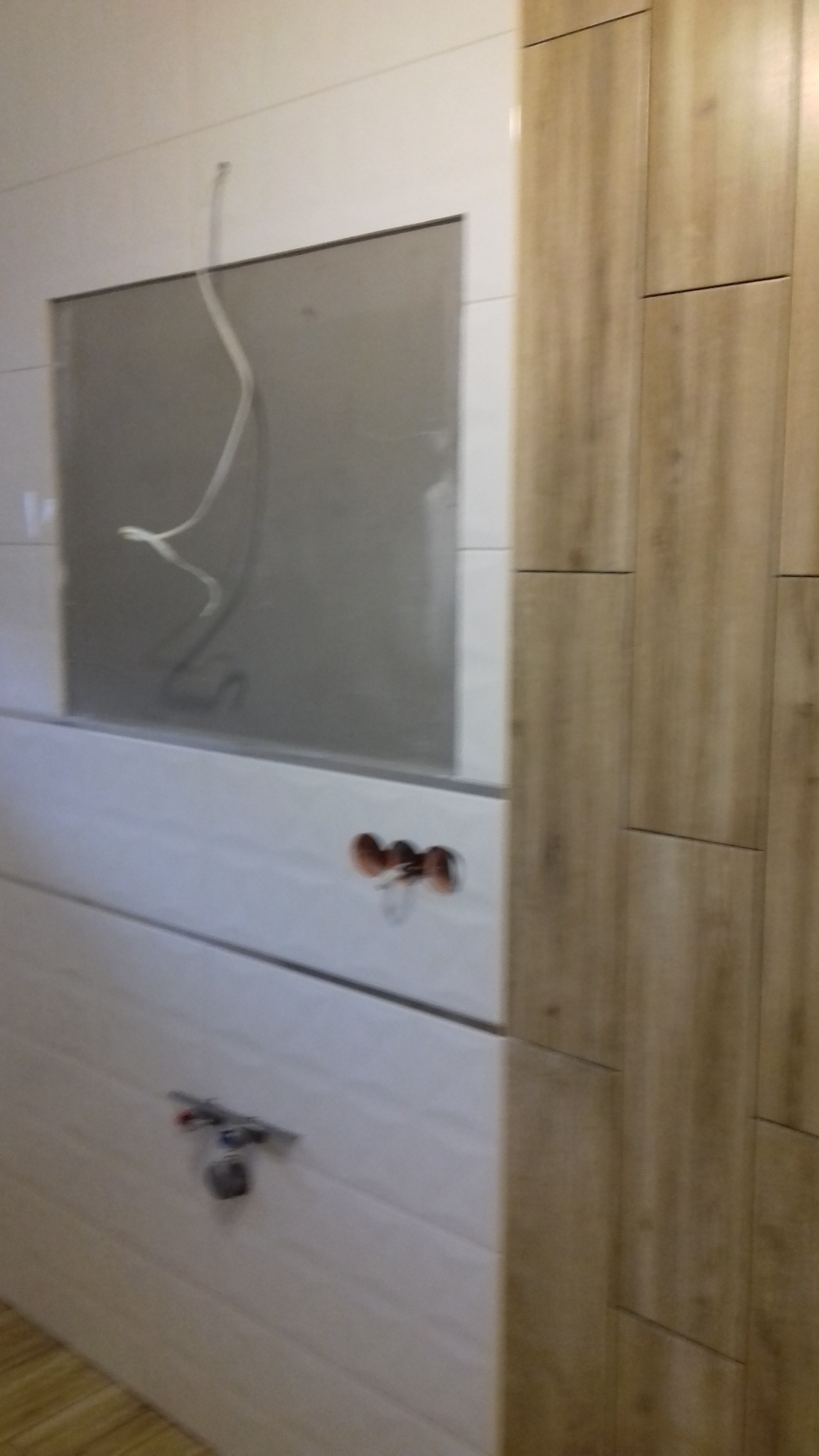 Wnętrze łazienki w trakcie wykańczania: białe płytki imitujące drewno z wyciętym otworem, widoczne przewody, oraz pionowy fragment ściany wyłożony płytkami w odcieniach beżu i brązu, również...