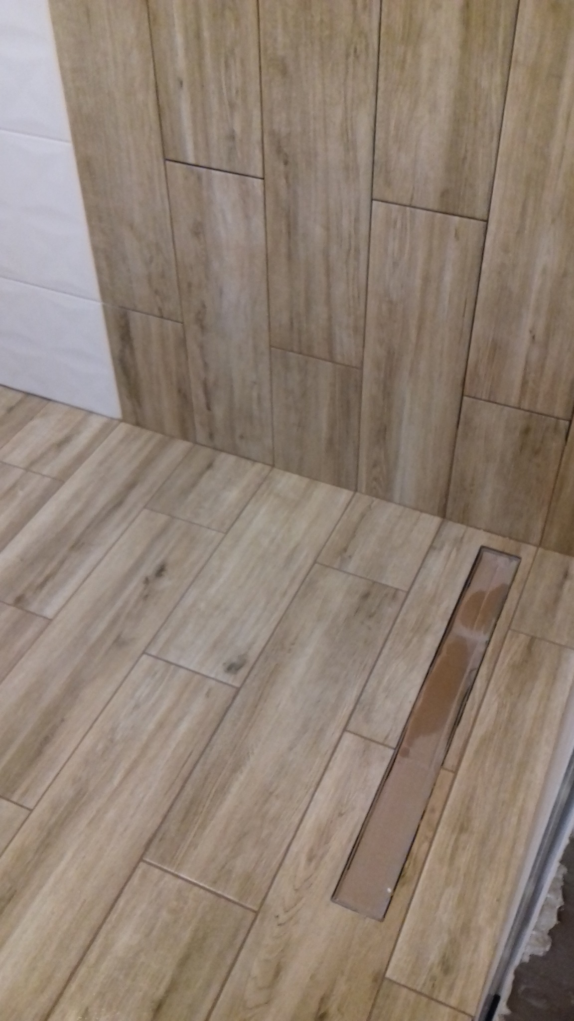 Ukończona łazienka z podłogą i ścianami wyłożonymi płytkami imitującymi drewno, z odpływem liniowym w podłodze.