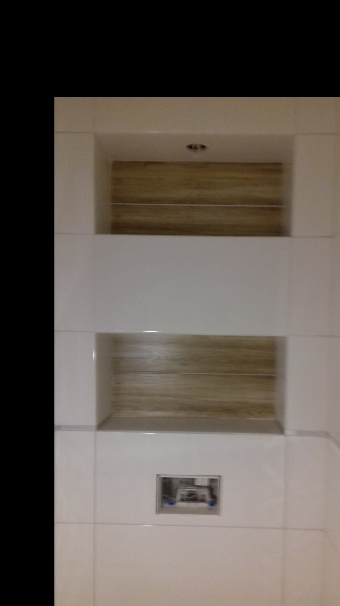 Białe płytki w łazience z dwoma wnękami wyłożonymi panelami imitującymi drewno; widoczna instalacja podtynkowa.