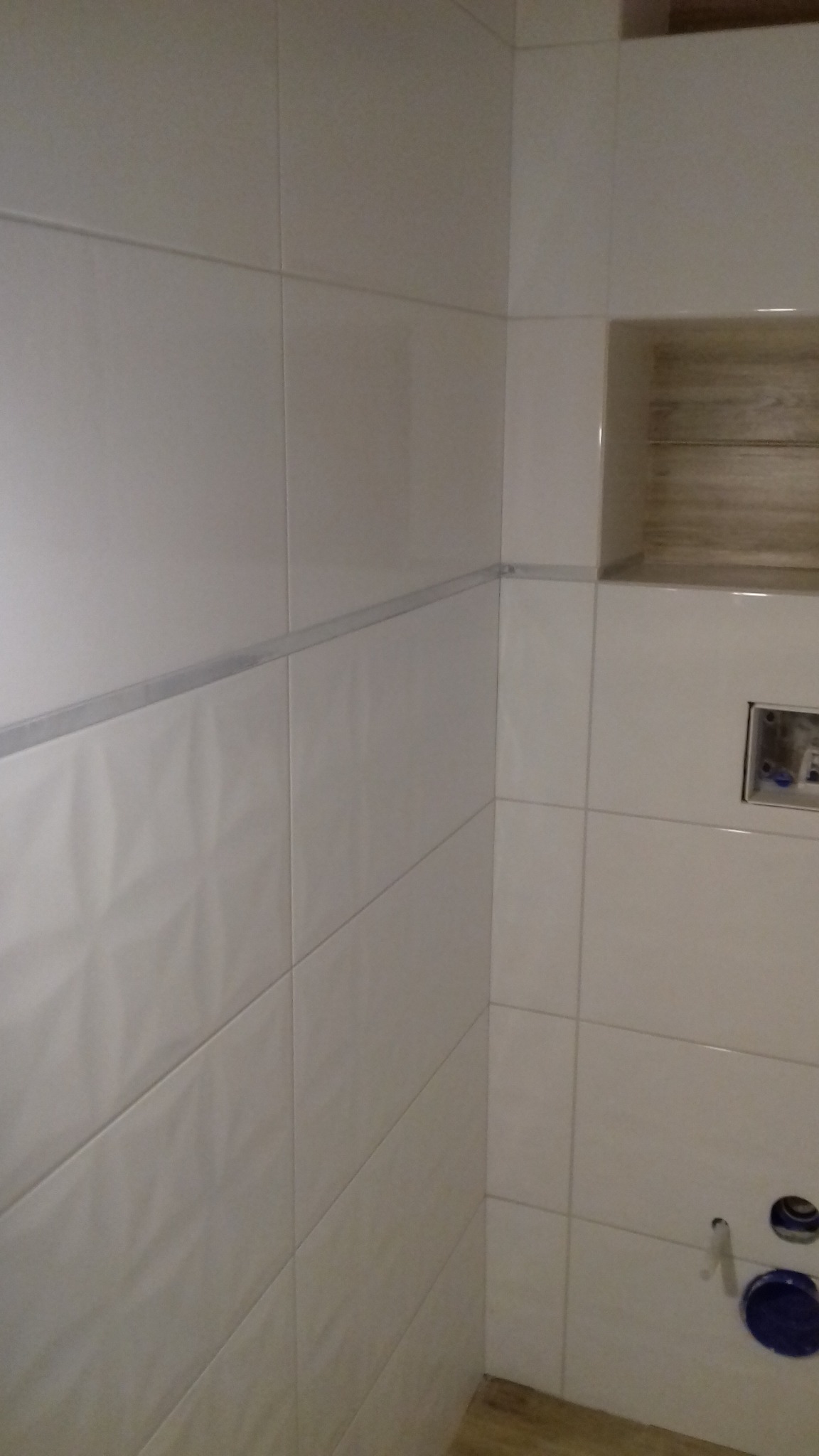 Ściana łazienki wyłożona białymi, błyszczącymi płytkami z dekoracyjnym pasem i wnęką z drewnopodobnym wykończeniem, widoczne elementy instalacji hydraulicznej.