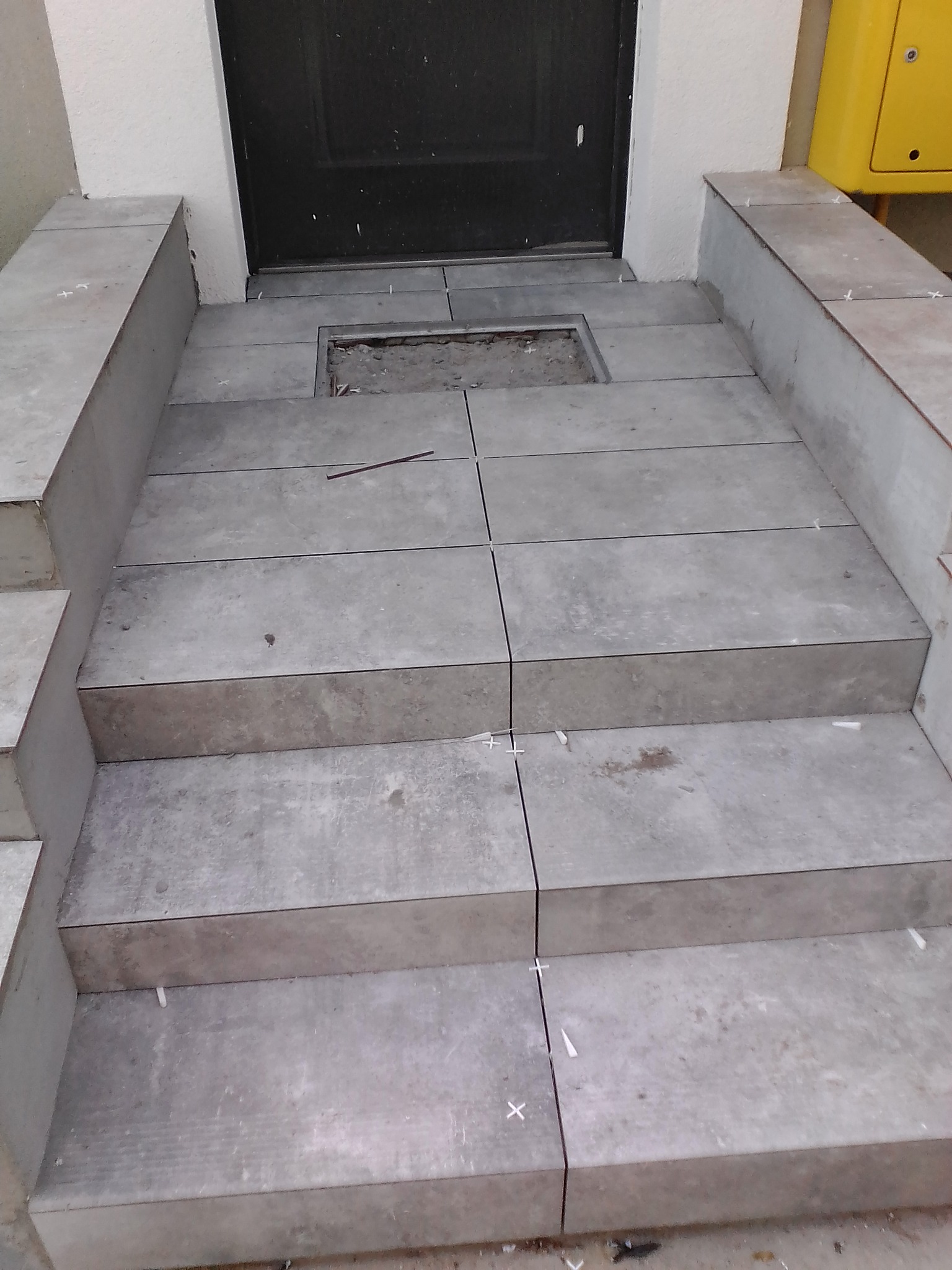 Schody wejściowe do budynku w trakcie układania szarych płytek, widoczne krzyżyki dystansowe i niezabezpieczony otwór rewizyjny.