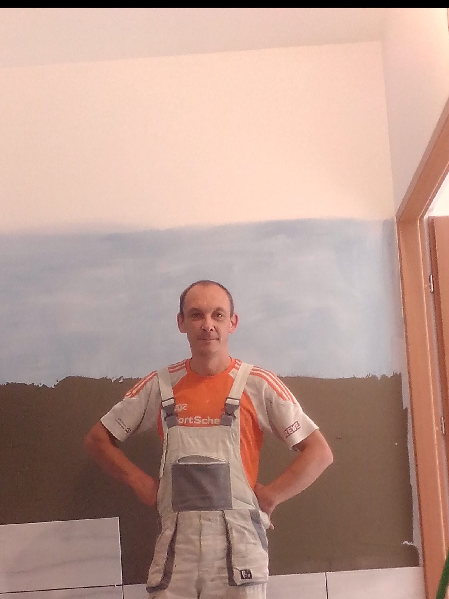 Mężczyzna w roboczym kombinezonie stoi na tle ściany z częściowo położonymi białymi płytkami i przygotowaną zaprawą, widoczne niedokończone malowanie górnej części ściany na niebiesko.