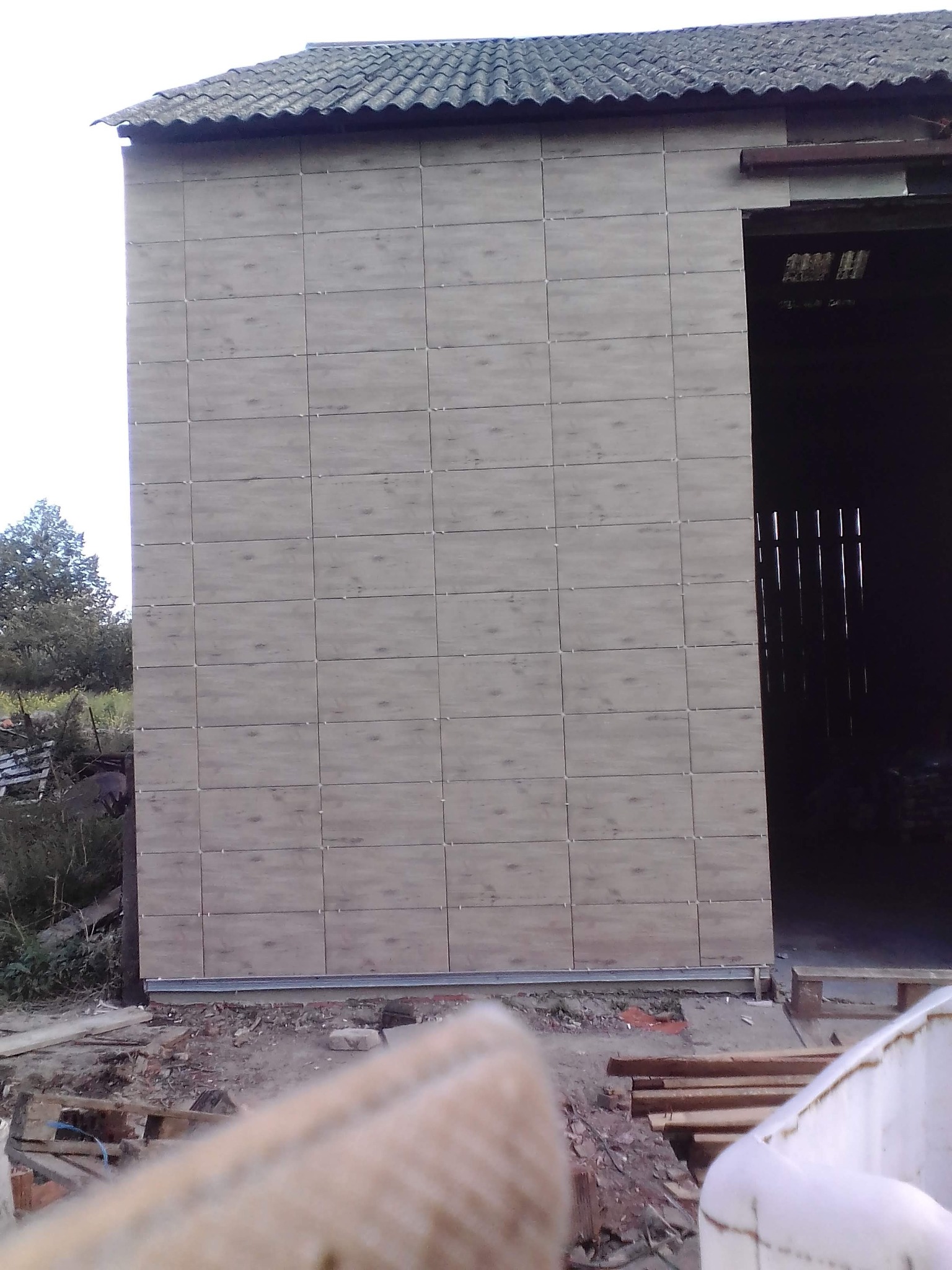 Elewacja budynku gospodarczego wykończona płytami imitującymi drewno, widoczne niedokończone prace ziemne u podstawy ściany i fragment wnętrza budynku.