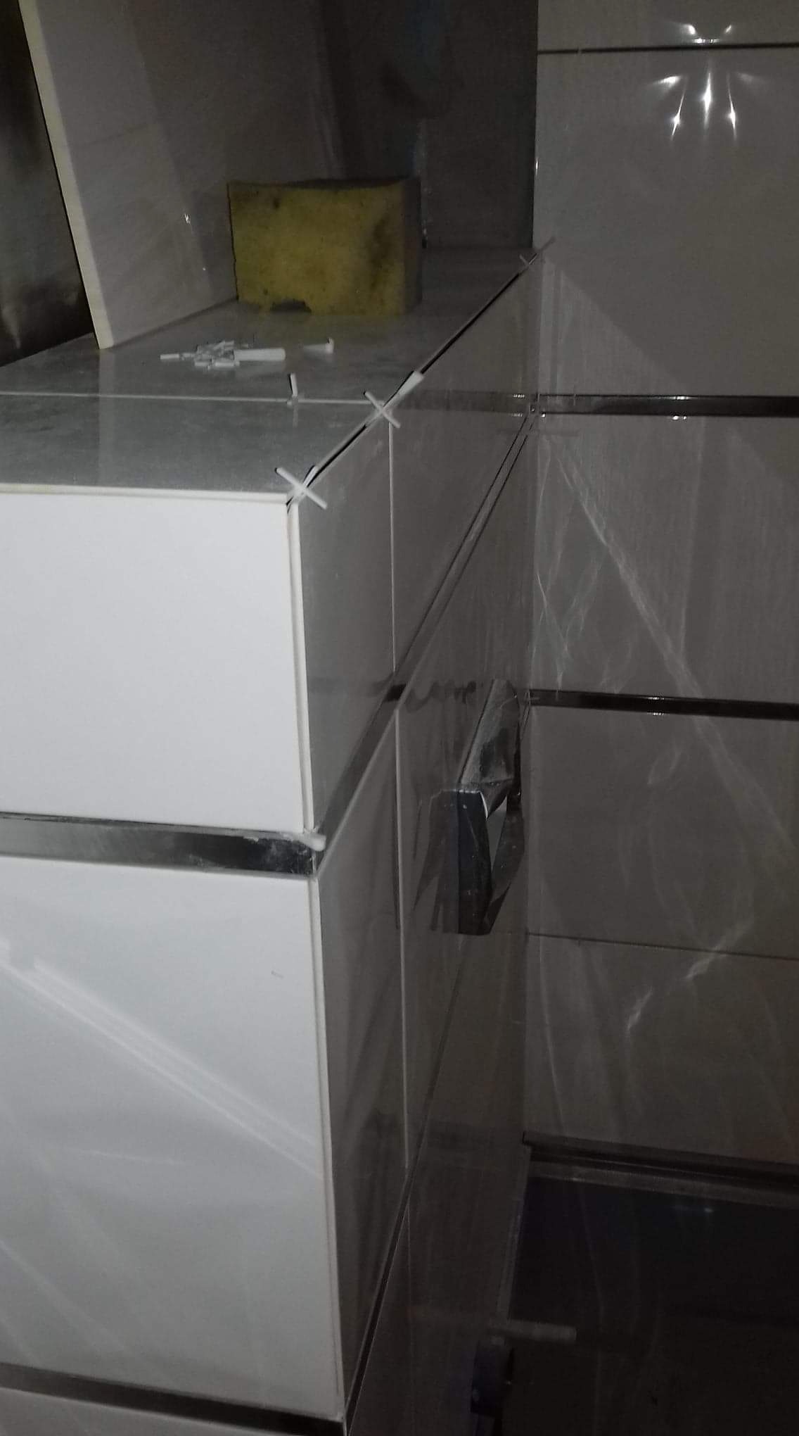 Układanie białych płytek z metalowymi listwami wykończeniowymi, krzyżyki dystansowe w spoinach, gąbka i strzykawki do fugowania na blacie, w tle folia zabezpieczająca.