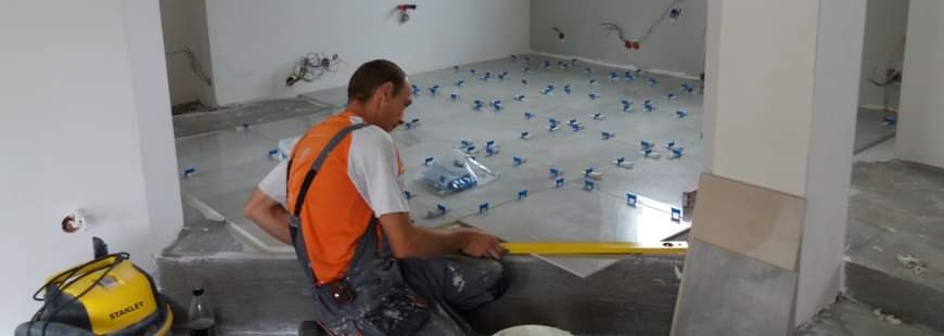 Mężczyzna w roboczym kombinezonie i pomarańczowej koszulce układa szare płytki na podłodze, używając poziomicy i plastikowych krzyżyków dystansowych; w tle widoczne instalacje elektryczne...