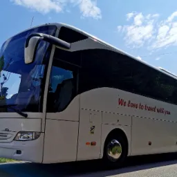 Biały autokar Mercedes z napisem 'We love to travel with you' na tle błękitnego nieba z chmurami.