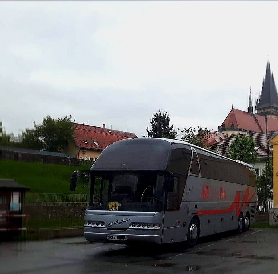 Szary autokar z napisem 'Exclusiv' na tle zabudowań miejskich z kościołem w tle, widoczny znak przewozu dzieci.