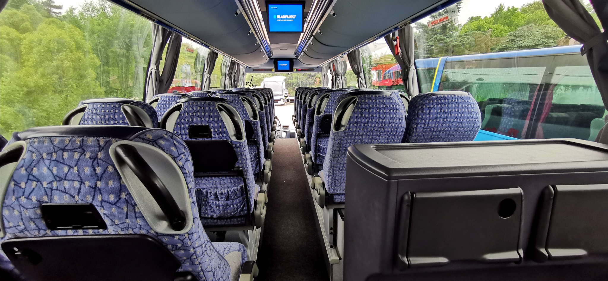 Wnętrze autobusu turystycznego z rzędami foteli obitych niebieską tkaniną we wzory, widoczne monitory Blaupunkt pod sufitem i zieleń za oknami.