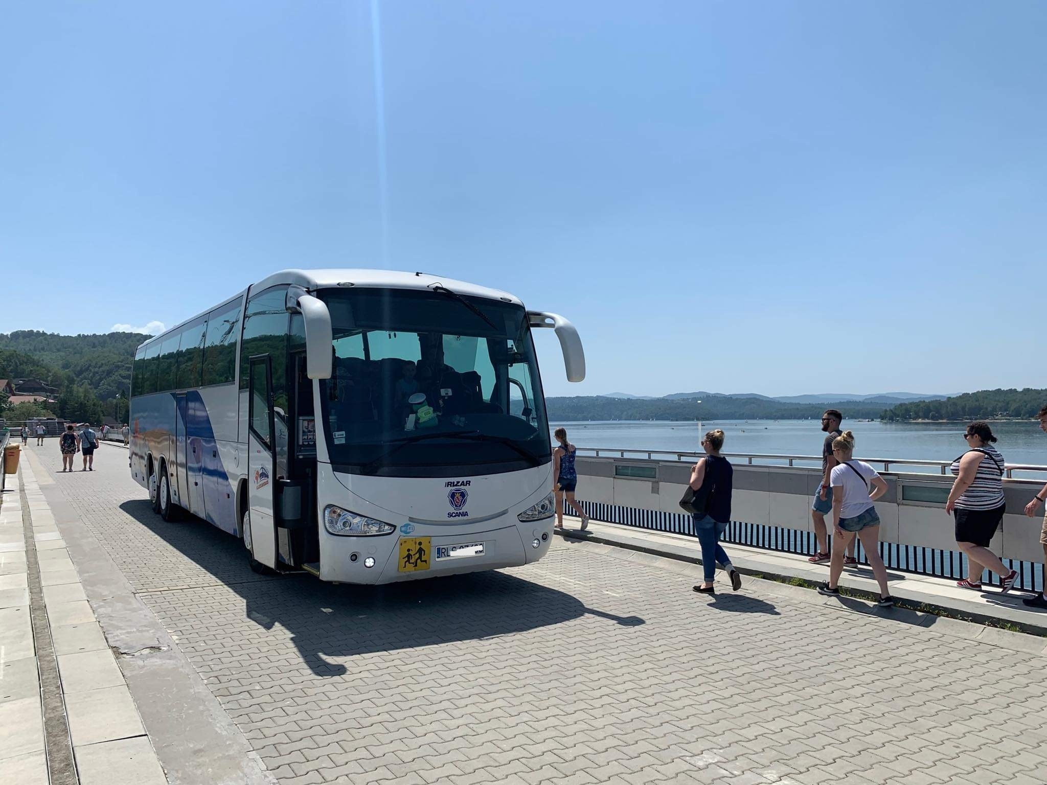 Biały autokar marki Irizar Scania z naklejką przewozu dzieci na tle jeziora i grupy ludzi w słoneczny dzień.