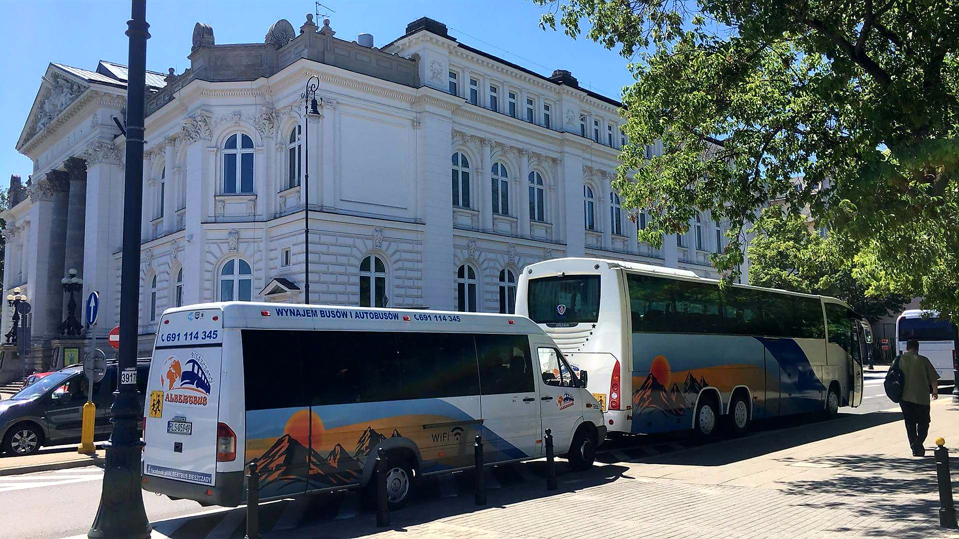 Dwa busy i autobus zaparkowane przy chodniku, z dekoracyjnym motywem górskim i zachodzącego słońca na boku, na tle klasycystycznego budynku z kolumnami.