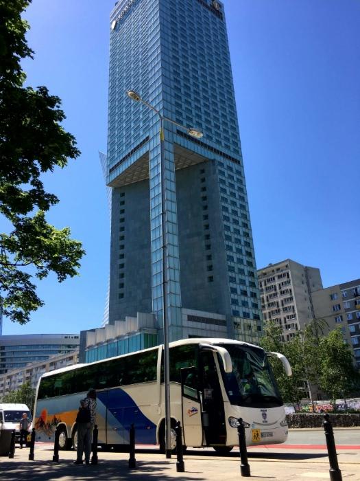 Autokar turystyczny z niebiesko-żółtym motywem graficznym na tle szklanego wieżowca w słoneczny dzień.