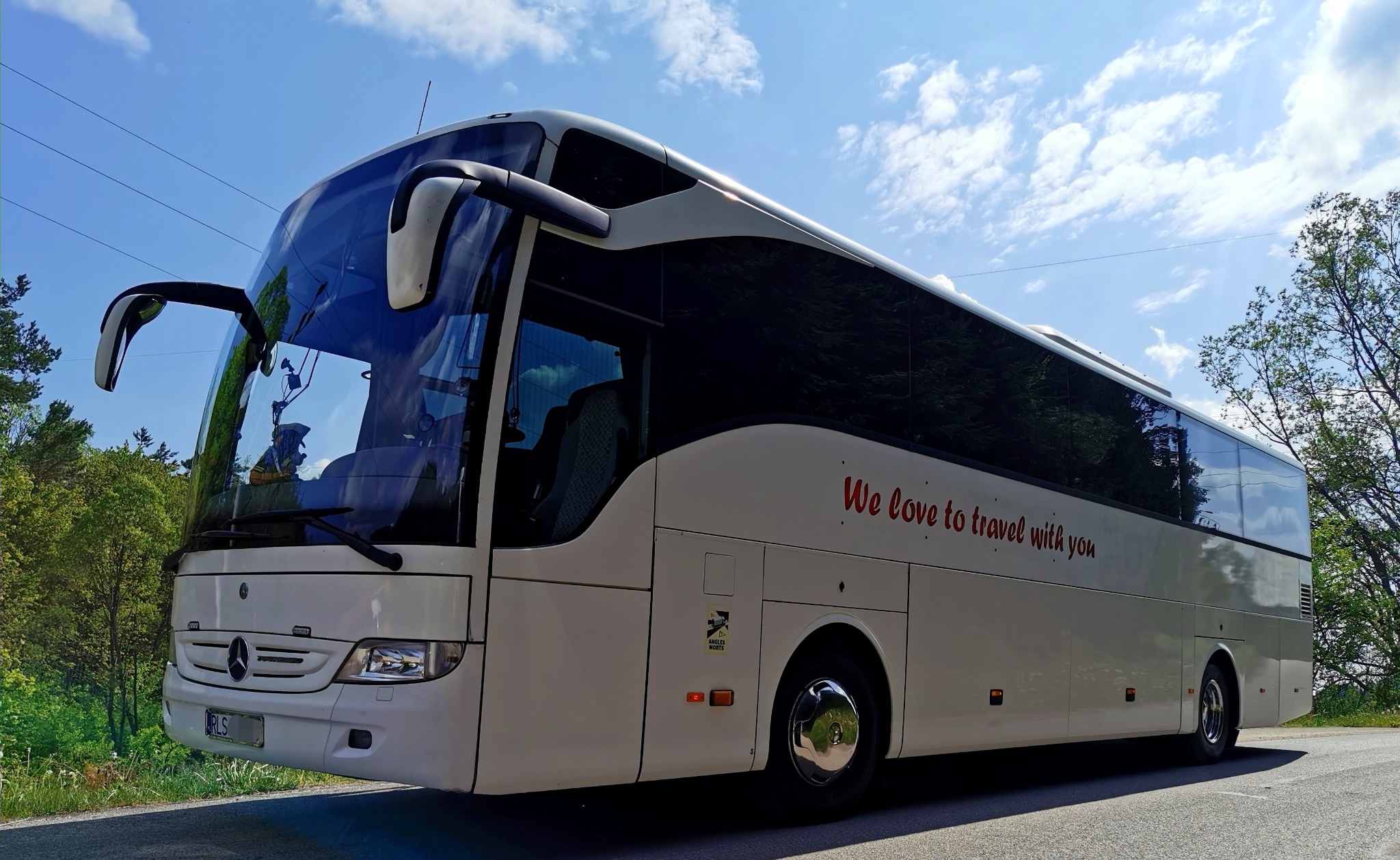 Biały autokar Mercedes z napisem 'We love to travel with you' na tle błękitnego nieba z chmurami.