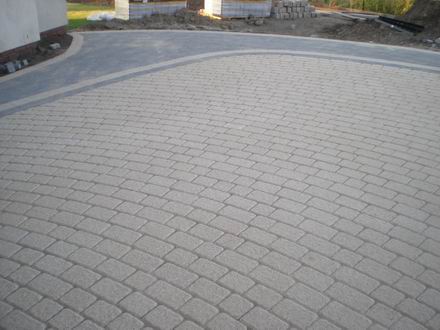 Ułożona kostka brukowa w odcieniach szarości, układana w łukowaty wzór, z widocznym obrzeżem z ciemniejszej kostki. Na obrzeżach widać palety z kostką brukową.