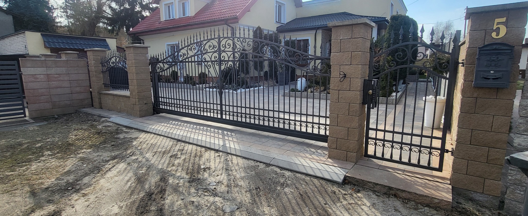 Kuta brama wjazdowa i furtka z kamiennymi słupkami, numer domu '5' i skrzynka pocztowa, kostka brukowa przed posesją.