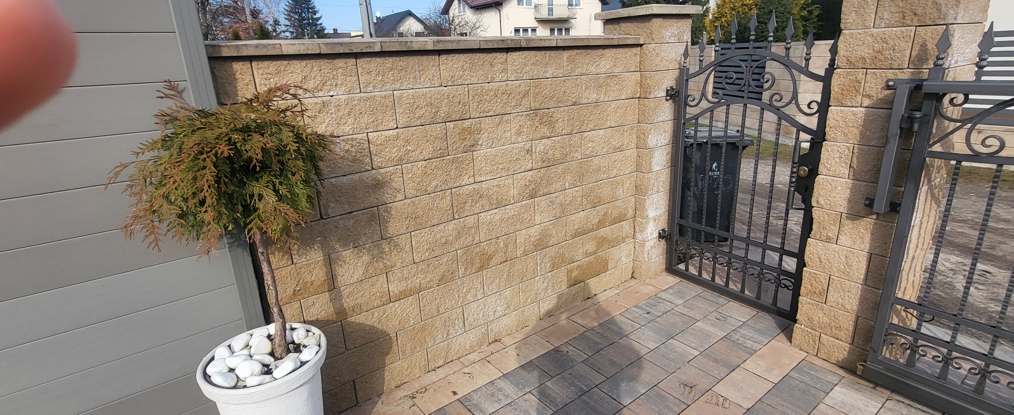 Ogrodzenie z bloczków betonowych z metalową bramą, obok tuja w donicy z białymi kamieniami, kostka brukowa na podjeździe.
