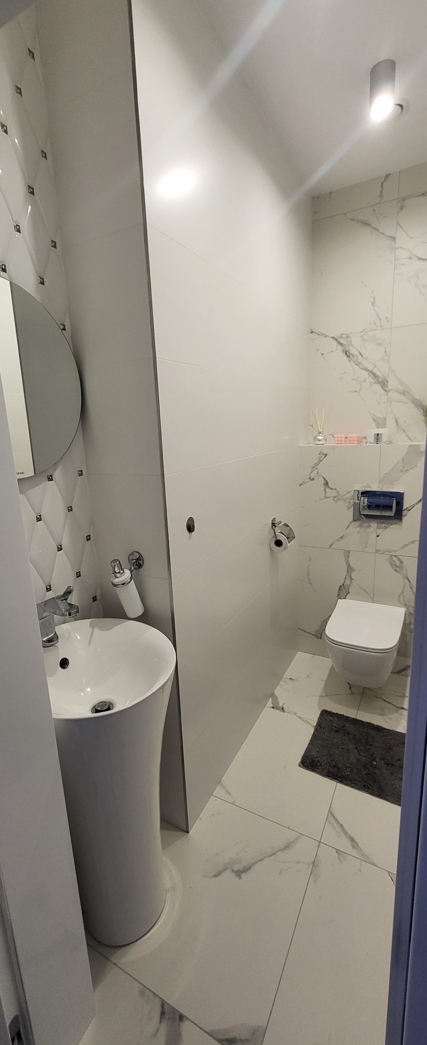 Nowoczesna łazienka z marmurowymi płytkami, owalnym lustrem i wolnostojącą umywalką na postumencie, oświetlona punktowym światłem.