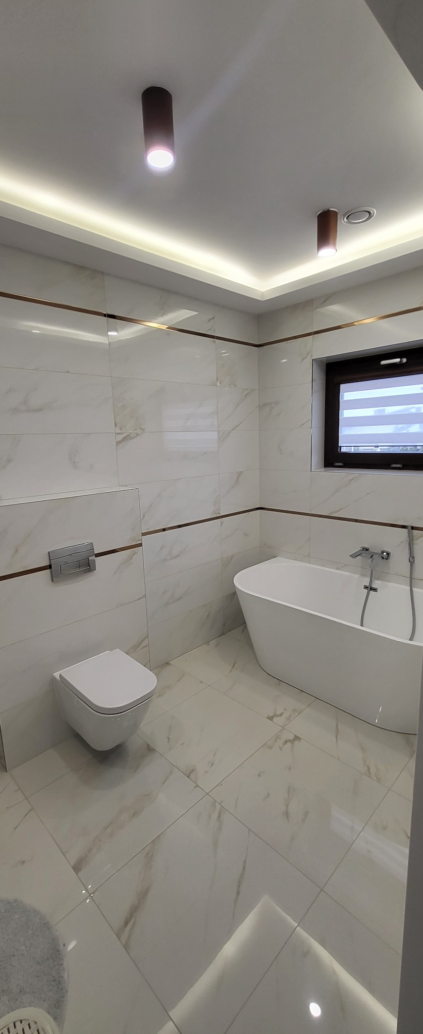 Nowoczesna łazienka z białymi marmurkowymi płytkami, podwieszaną toaletą, wolnostojącą wanną i oknem z żaluzjami, oświetlona punktowymi lampami i listwą LED.