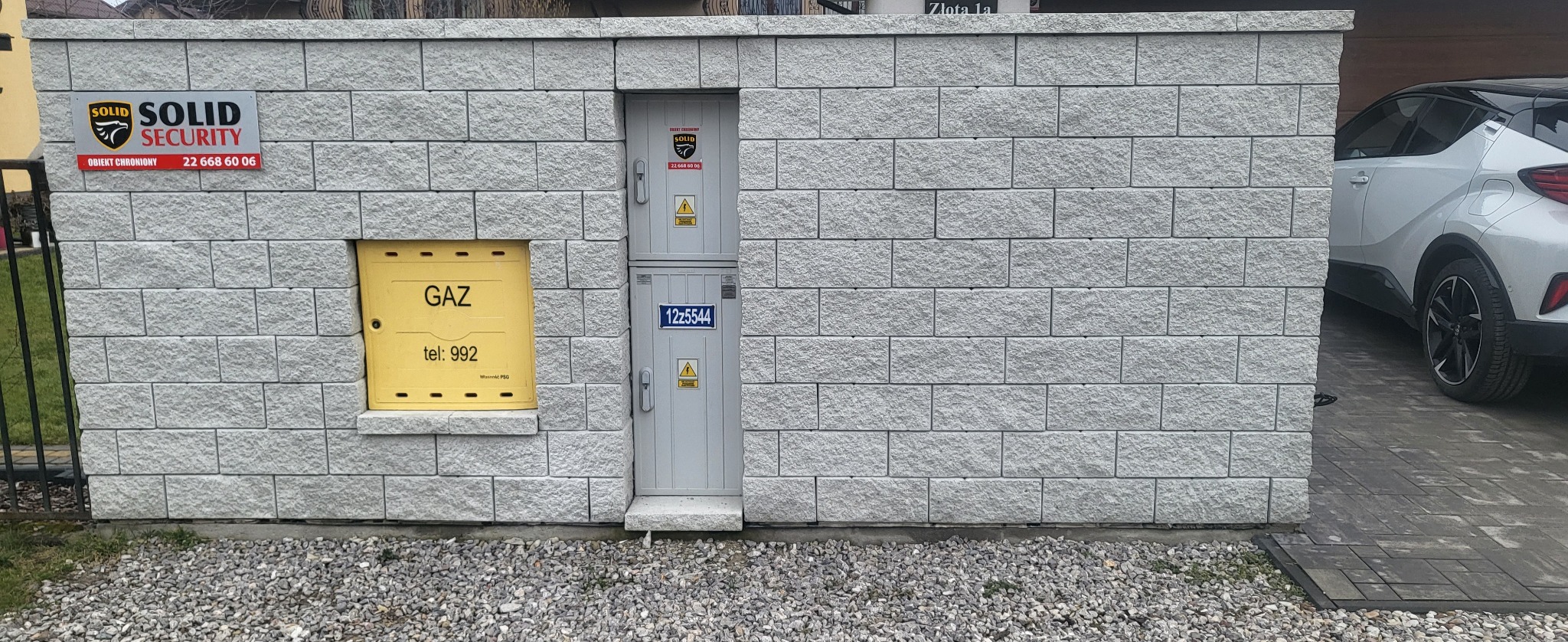 Szary mur z bloków betonowych z żółtą szafką gazową i szarą szafką elektryczną, na tle zaparkowany biały samochód.