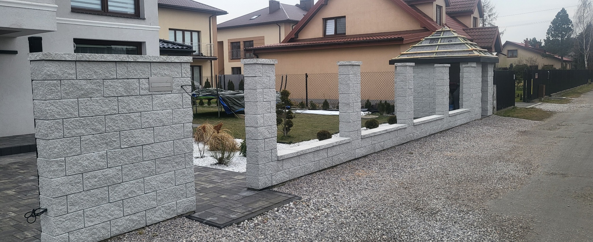 Nowoczesne ogrodzenie z szarej kostki betonowej z filarami i małą altaną, widok na posesję z trampoliną i sąsiednie domy.