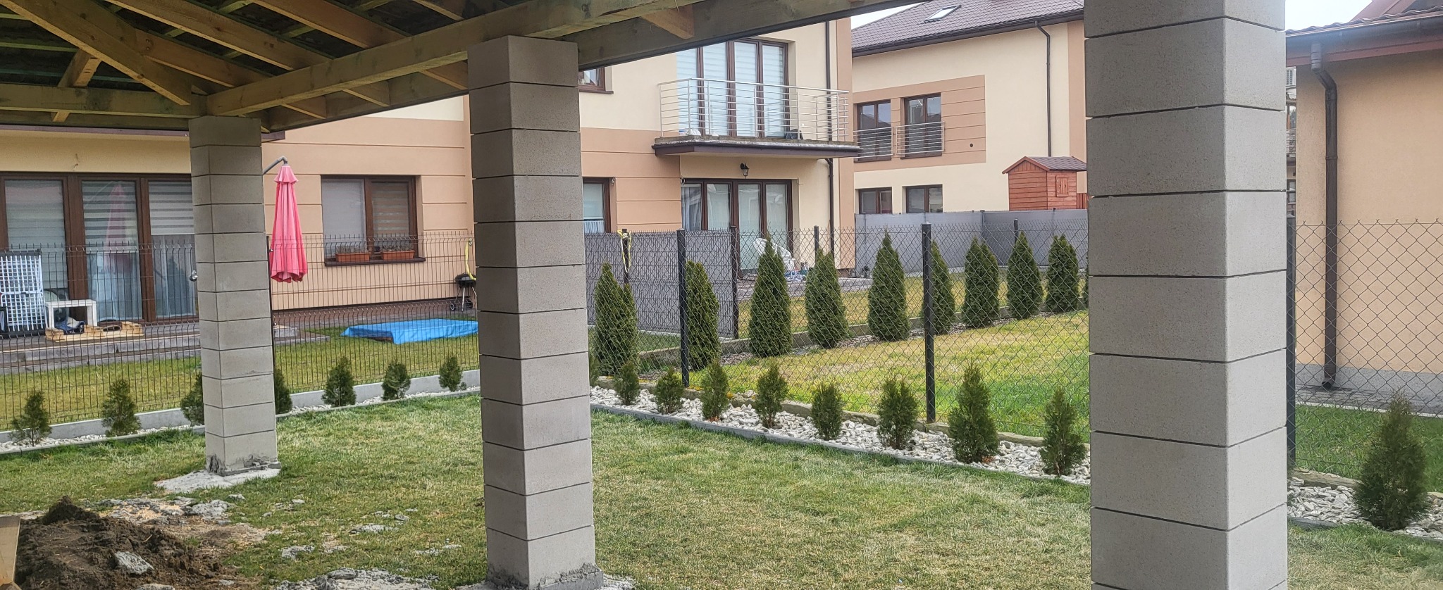 Wznoszenie zadaszenia altany z betonowych bloczków, widoczny fragment ogrodzenia panelowego i nasadzenia z tujek w ogrodzie.