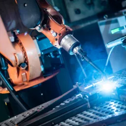 Pomarańczowy robot spawalniczy w trakcie pracy: intensywne niebieskie światło spawania oświetla metalowe elementy mocowane zaciskami na perforowanym stole.