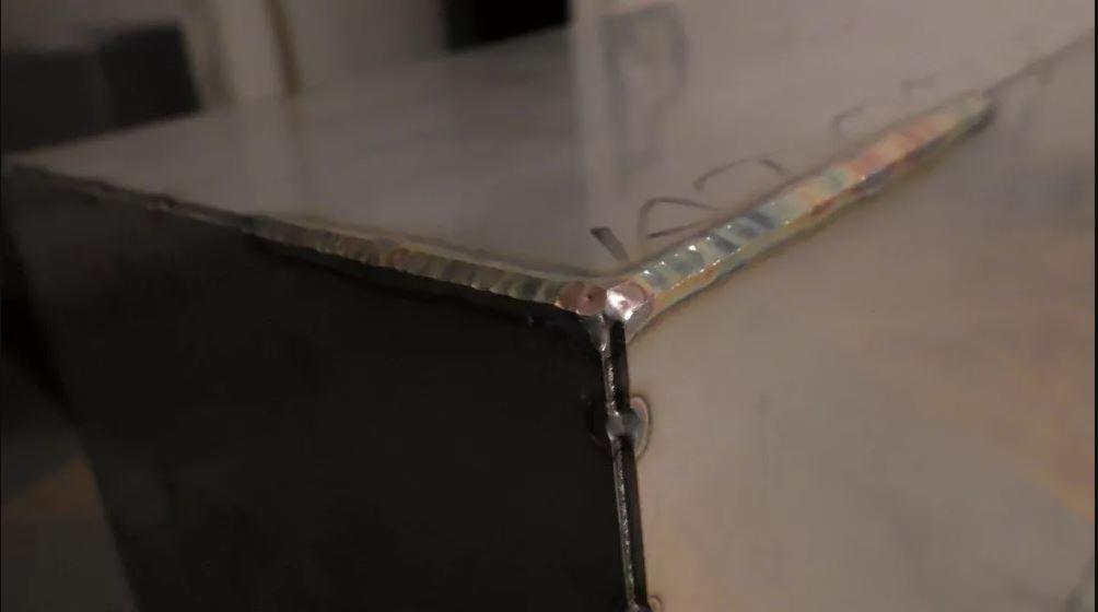 Spoina narożna łącząca dwie metalowe płyty, widoczne ślady po procesie łączenia, różnobarwne przebarwienia termiczne na spoinie.