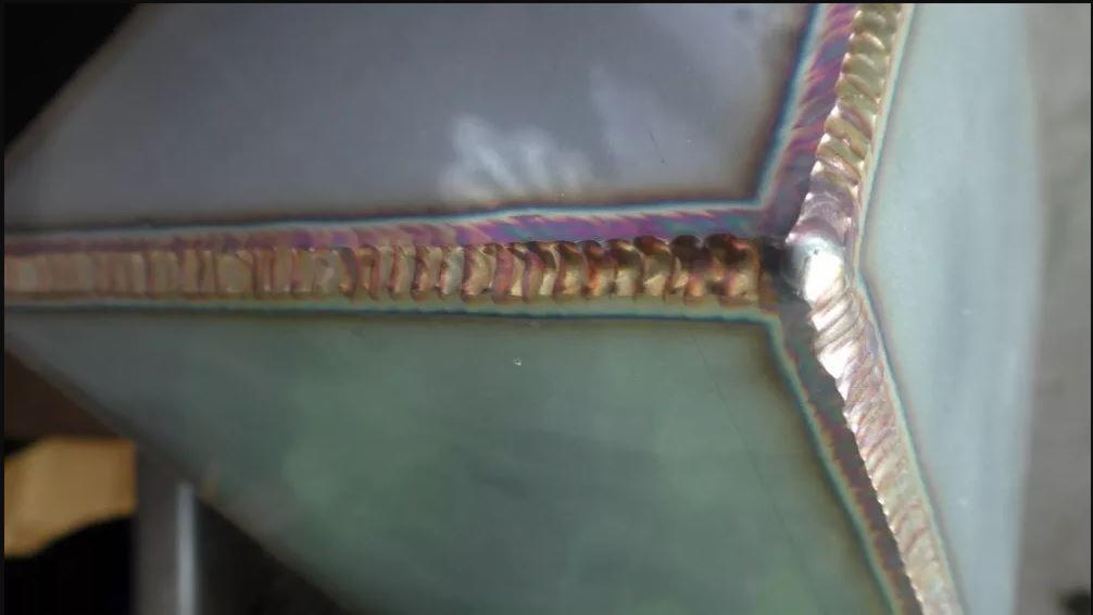 Szczegółowe ujęcie spoiny łączącej metalowe elementy pod kątem, widoczne tęczowe przebarwienia termiczne na spoinie.