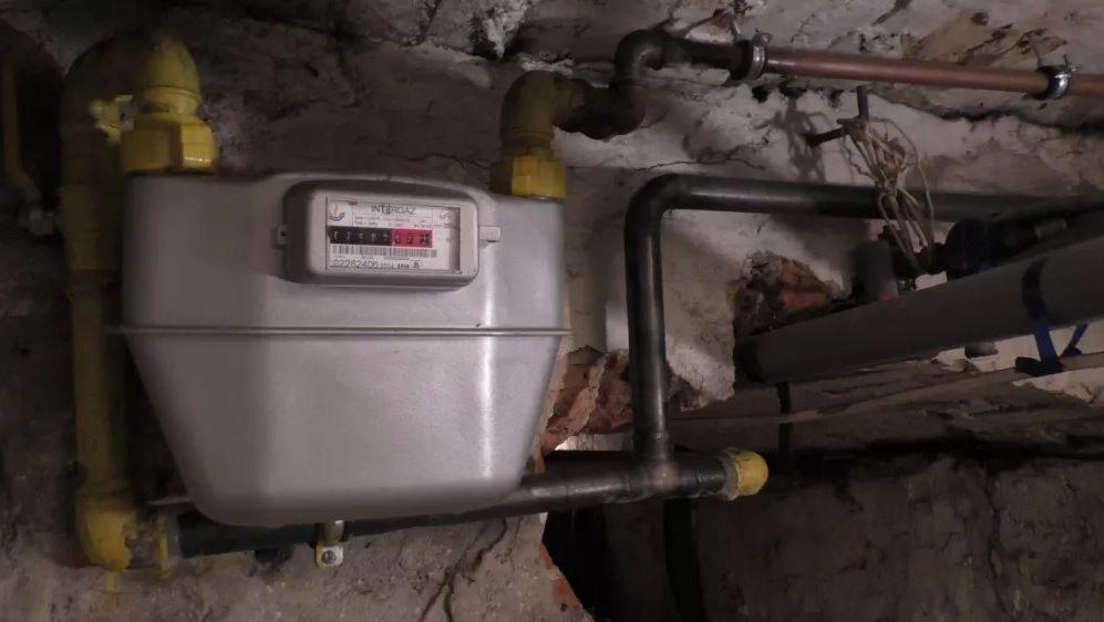 Szary gazomierz Intergaz z żółtymi rurami w piwnicy z kamiennymi ścianami i różnymi rurami.