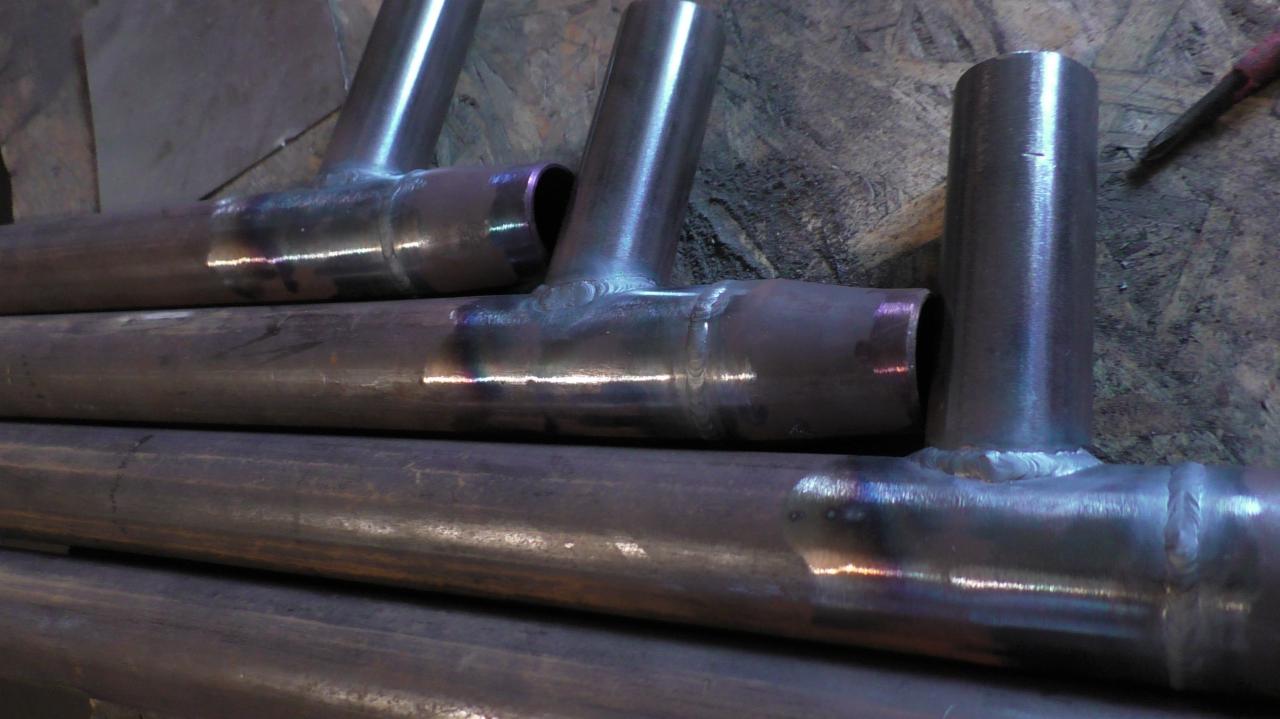 Metalowe rury spawane w kształcie litery T, widoczne ślady po spawaniu z opalizującymi przebarwieniami, ułożone na drewnianej powierzchni.