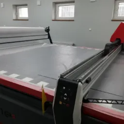 Automatyczna krojownia z rolką materiału, widoczne okna w tle, maszyna Unicut One S.