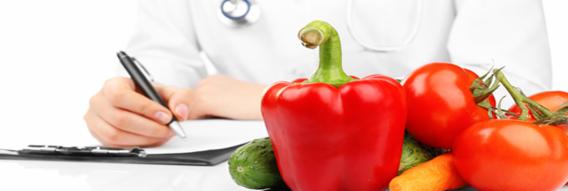 Osoba w białym fartuchu z stetoskopem na szyi pisze na podkładce, obok leżą świeże warzywa: czerwona papryka, pomidory, ogórek i marchewka.