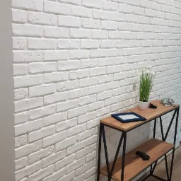 Ściana wykończona białymi cegłami dekoracyjnymi, z drewnianym stolikiem konsolą z czarnymi metalowymi nogami, na którym stoją ramka, roślina w doniczce i okulary.