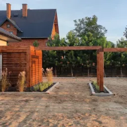 Drewniana konstrukcja altany w trakcie budowy, widoczne słupy i belki, świeżo posadzone rośliny ozdobne, teren przygotowany pod dalsze prace, dom w tle.