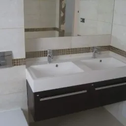 Nowoczesna łazienka z podwójną umywalką nablatową na szafce w kolorze ciemnego brązu, wykończona beżowymi płytkami z dekoracyjnym pasem mozaiki, oraz chromowaną armaturą.