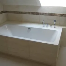 Wanna obudowana jasnymi płytkami, z dekoracyjnym pasem mozaiki na ścianie, bateria wannowa trzyotworowa, oświetlenie z okna dachowego.