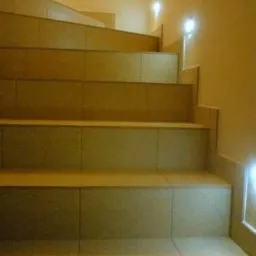 Schody wewnętrzne wyłożone płytkami ceramicznymi w odcieniach beżu, z dyskretnym oświetleniem punktowym na ścianie i stopniach.