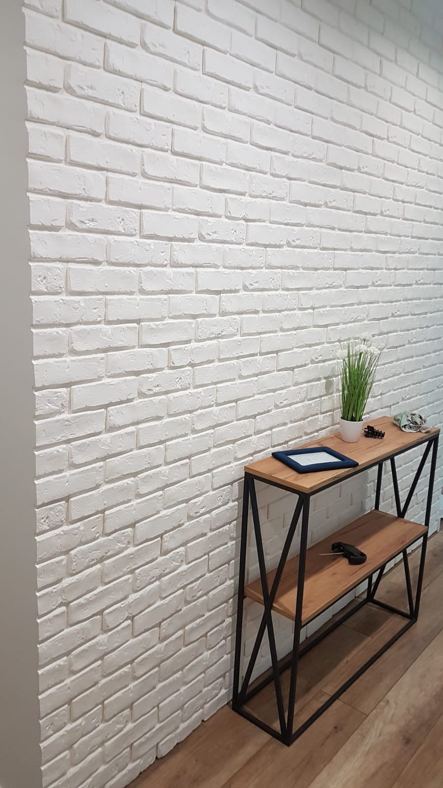 Ściana wykończona białymi cegłami dekoracyjnymi, z drewnianym stolikiem konsolą z czarnymi metalowymi nogami, na którym stoją ramka, roślina w doniczce i okulary.
