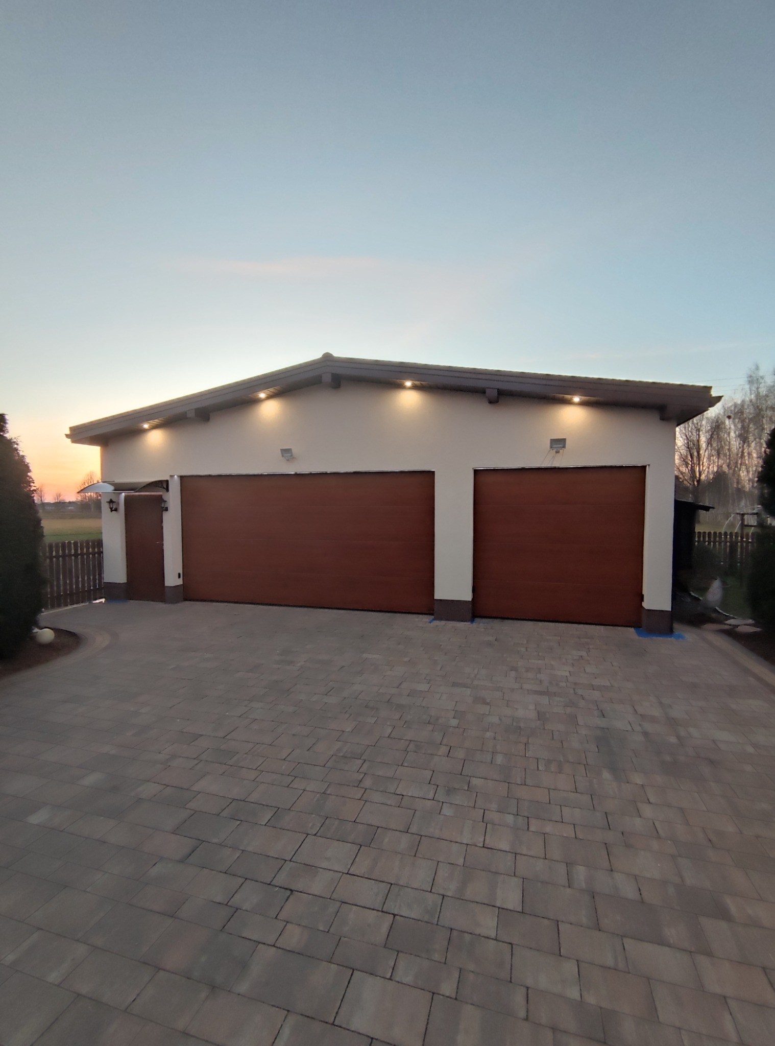 Nowoczesny garaż wolnostojący z dwoma brązowymi bramami segmentowymi i bocznym wejściem, oświetlony lampami zewnętrznymi, z kostki brukowej na podjeździe, sfotografowany o zmierzchu.