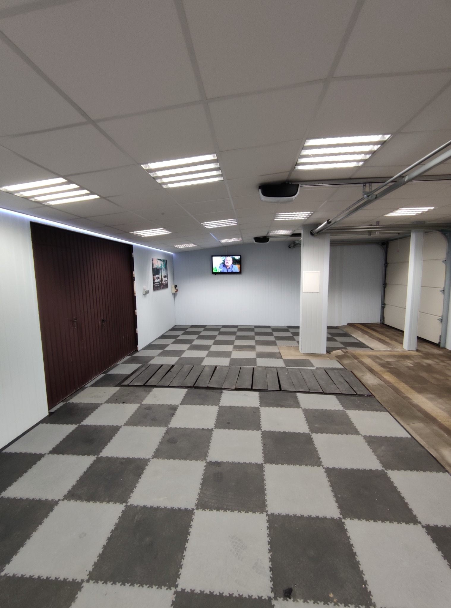 Wnętrze garażu z szachownicą na podłodze, brązowymi drzwiami garażowymi po lewej i białymi po prawej, oświetleniem sufitowym i telewizorem na ścianie.