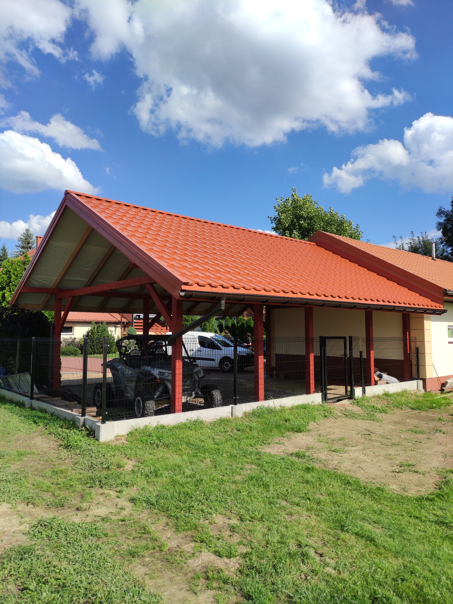 Zadaszenie z czerwoną dachówką, wsparte na drewnianych słupach, chroniące pojazdy ATV za czarnym ogrodzeniem panelowym.