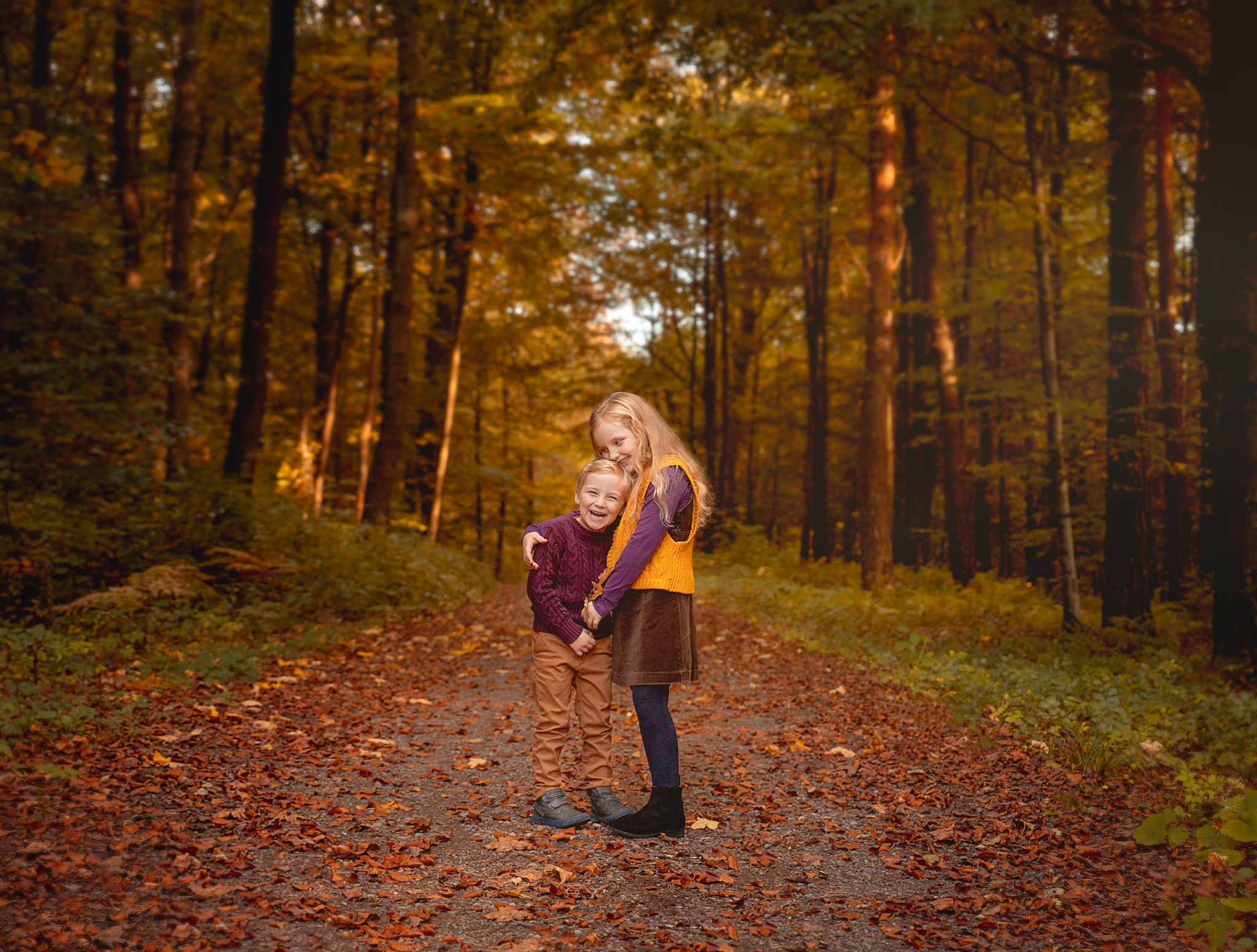 Uśmiechnięte rodzeństwo, dziewczynka obejmuje chłopca, stoją na ścieżce w jesiennym lesie, otoczeni opadłymi liśćmi.