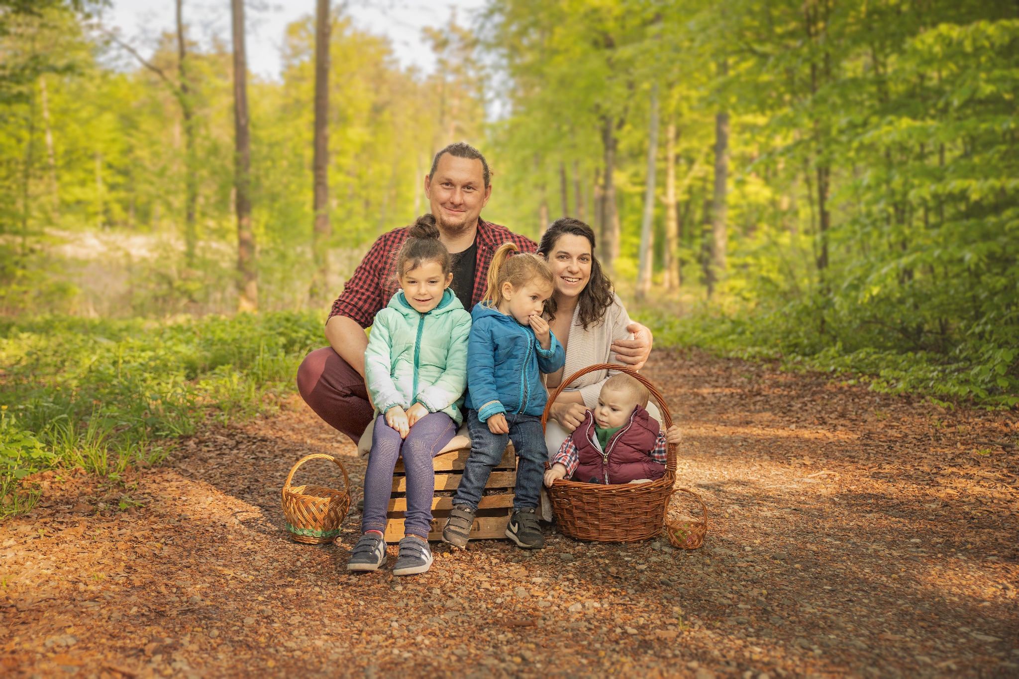 Rodzinne zdjęcie w lesie: rodzice z trójką dzieci siedzą na drewnianej skrzyni i w koszyku, otoczeni zielenią drzew i leśną ścieżką.