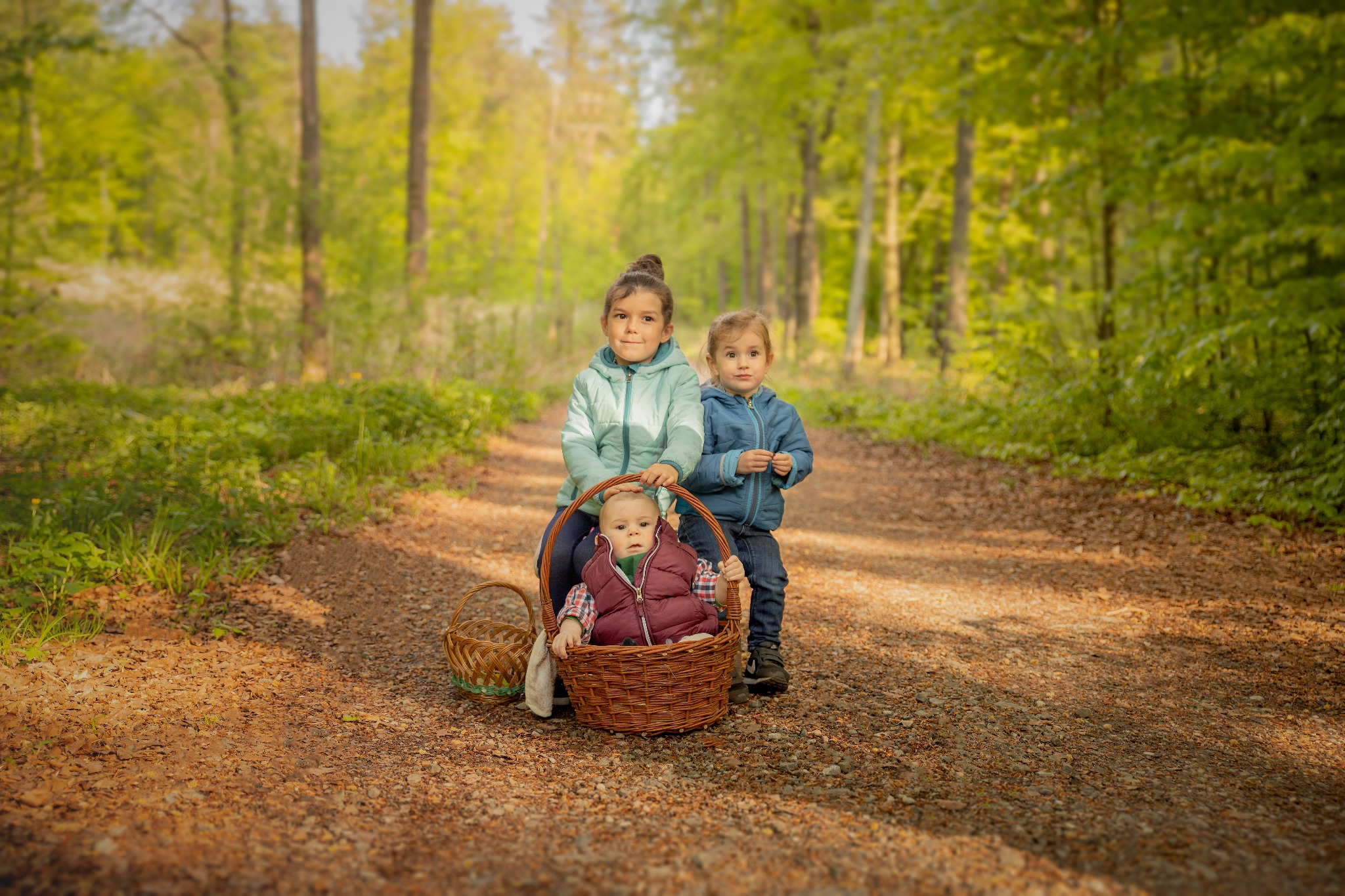 Trójka dzieci na leśnej ścieżce: niemowlę siedzi w wiklinowym koszu, a dwie starsze dziewczynki stoją za nim, w tle zielony las.