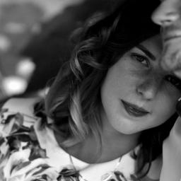 Karalina Kurhuzava - Portret kobiety z kręconymi włosami w kwiecistej sukience, opierającej głowę o ramię mężczyzny, czarno-białe zdjęcie z delikatnym uśmiechem.