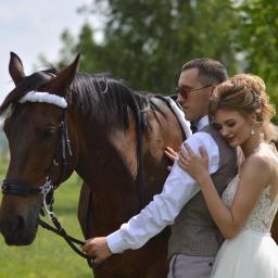 Karalina Kurhuzava - Para młoda w objęciach, obok brązowy koń z białym ozdobnym paskiem na głowie, w tle zielone drzewa i łąka.
