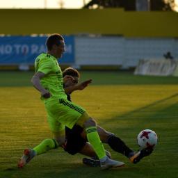 Karalina Kurhuzava - Piłkarz w jaskrawozielonym stroju uderza piłkę stopą, drugi zawodnik próbuje go powstrzymać, w tle żółta trybuna i baner z napisem 'Say No To Racism'.