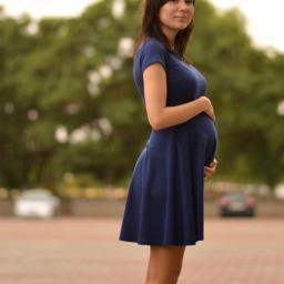 Karalina Kurhuzava - Kobieta w niebieskiej sukience ciążowej, profil, dłonie na brzuchu, zamazane tło miejskie z zielenią i padającym deszczem.