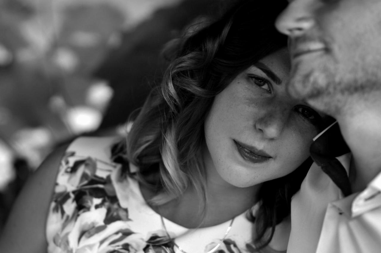 Portret kobiety z kręconymi włosami w kwiecistej sukience, opierającej głowę o ramię mężczyzny, czarno-białe zdjęcie z delikatnym uśmiechem.