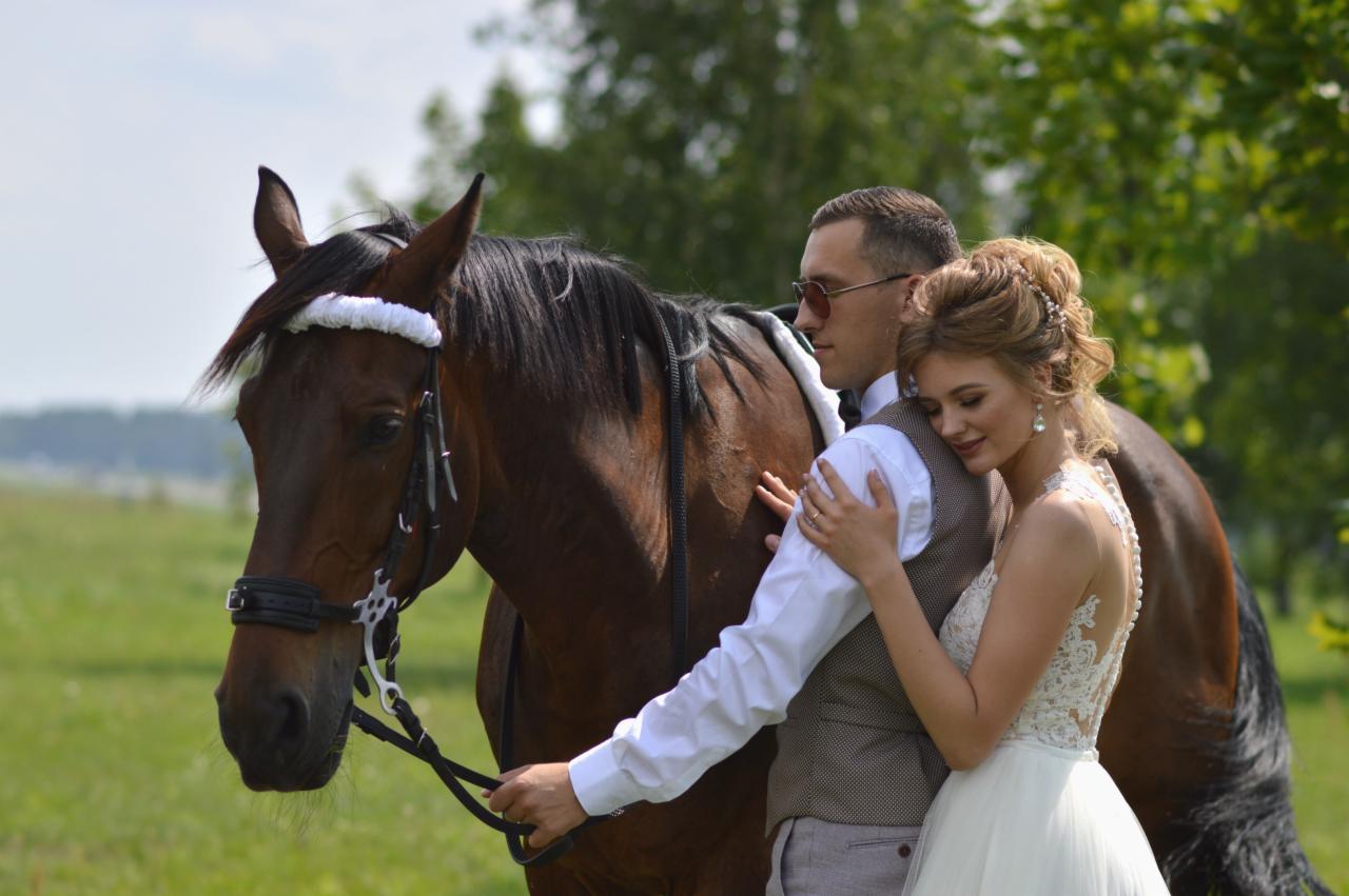 Para młoda w objęciach, obok brązowy koń z białym ozdobnym paskiem na głowie, w tle zielone drzewa i łąka.